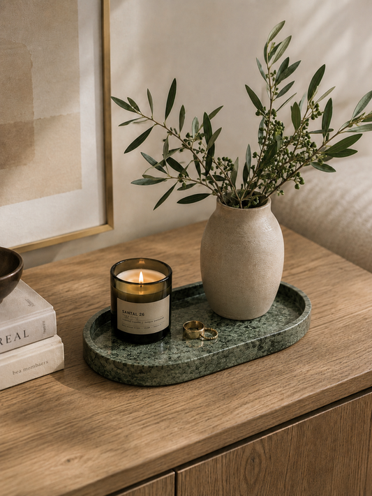 A styled interior scene showing a green marble tray holding a lit candle, a small vase with olive branches, and two rings on a wooden cabinet. A framed print and books are partially visible in the background.
