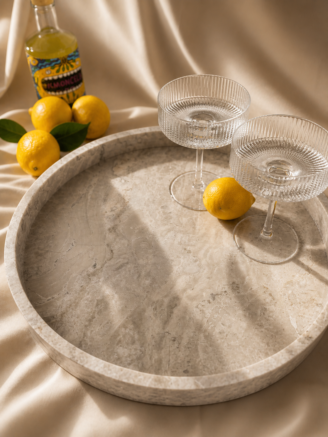 Round stone tray with two clear glasses, lemons, and a bottle on a beige fabric background